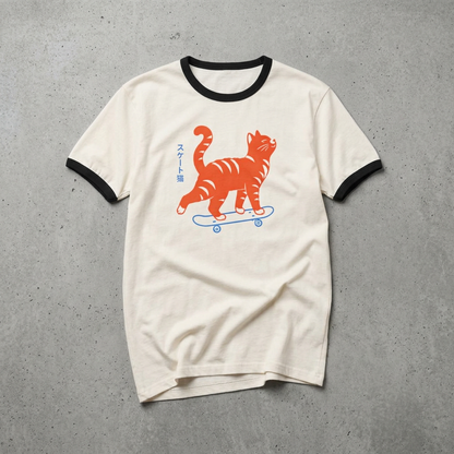 An off-white ringer t-shirt with black trim on the collar and sleeves, featuring a graphic of an orange tabby cat riding a blue skateboard with Japanese text on the front, laid flat on a gray concrete background.