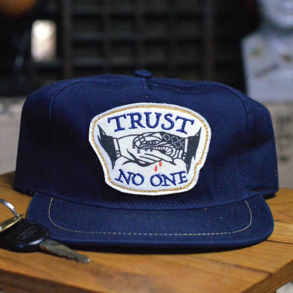 Blue cap with 'Trust No One' patch on a wooden surface