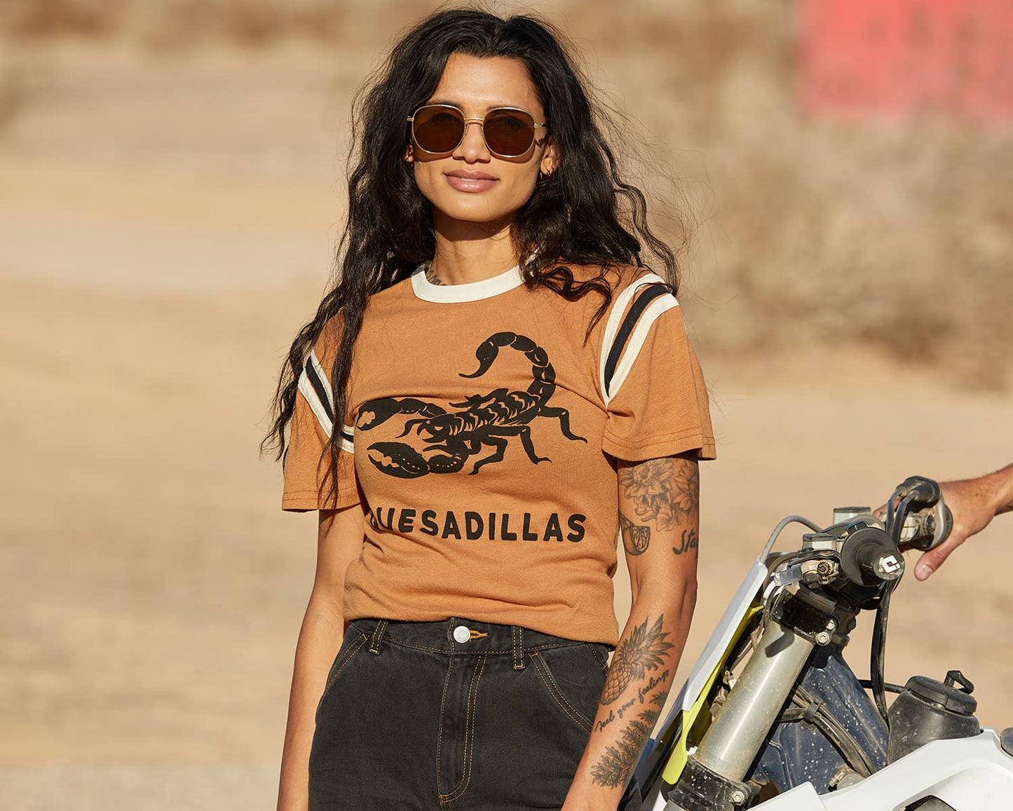 Woman wearing a brown t-shirt with a scorpion graphic and 'MEXICADILLAS' text, standing next to a motorcycle in a desert-like setting.