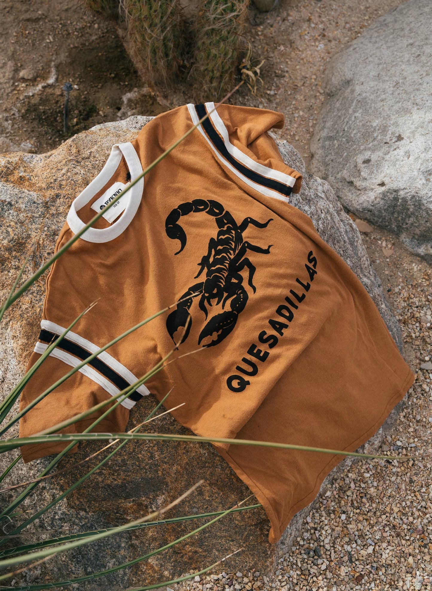 Brown shirt with black scorpion graphic and 'Quesadillas' text on a rocky surface