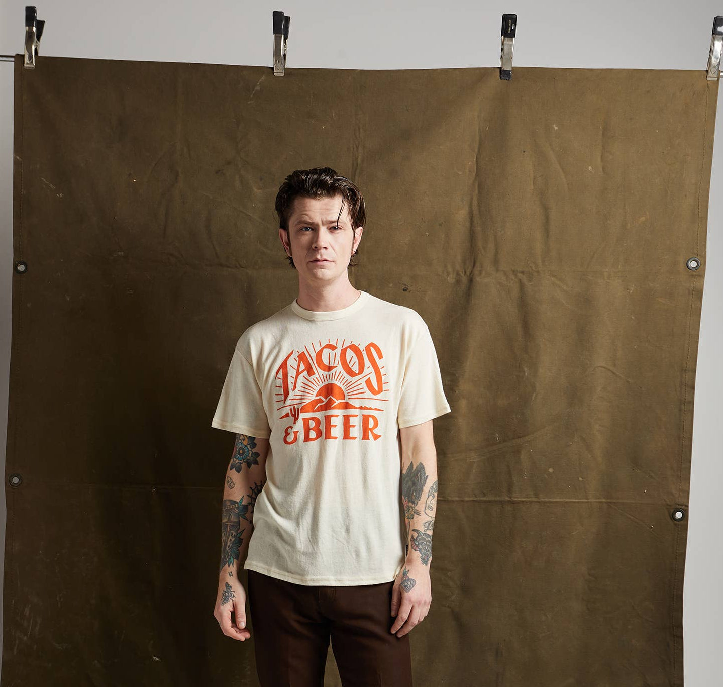 Person wearing a white t-shirt with red text and graphics, standing against a brown backdrop.