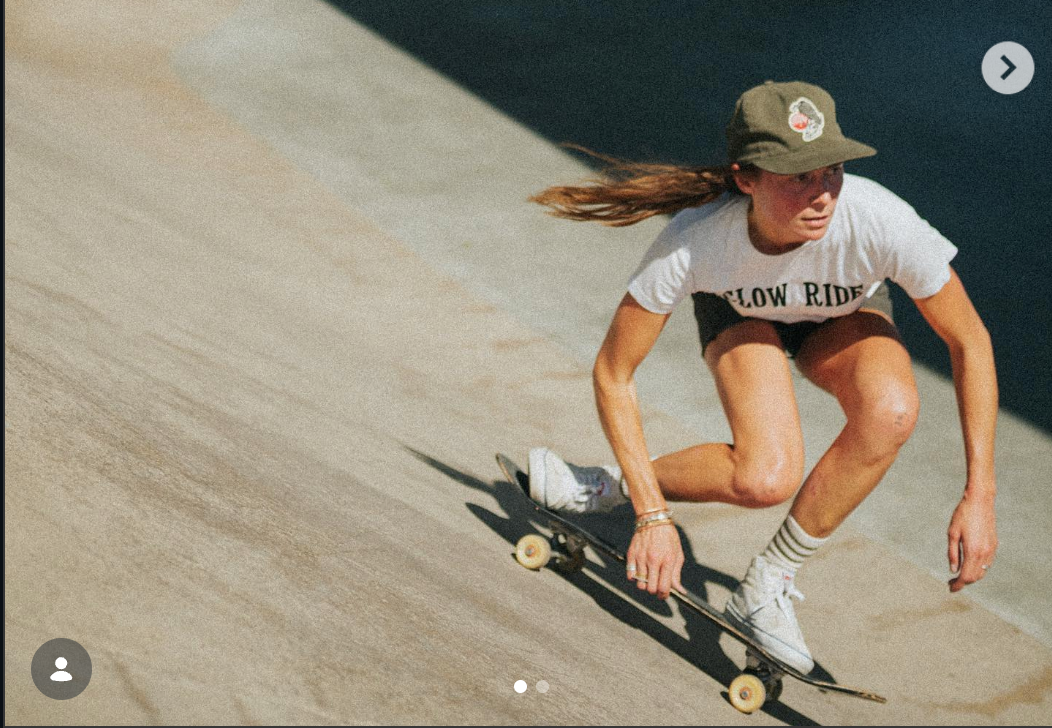 A woman skateboarding down a concrete ramp wearing a white graphic t-shirt, brown shorts, white sneakers, and a green cap, captured in a sunny outdoor setting.