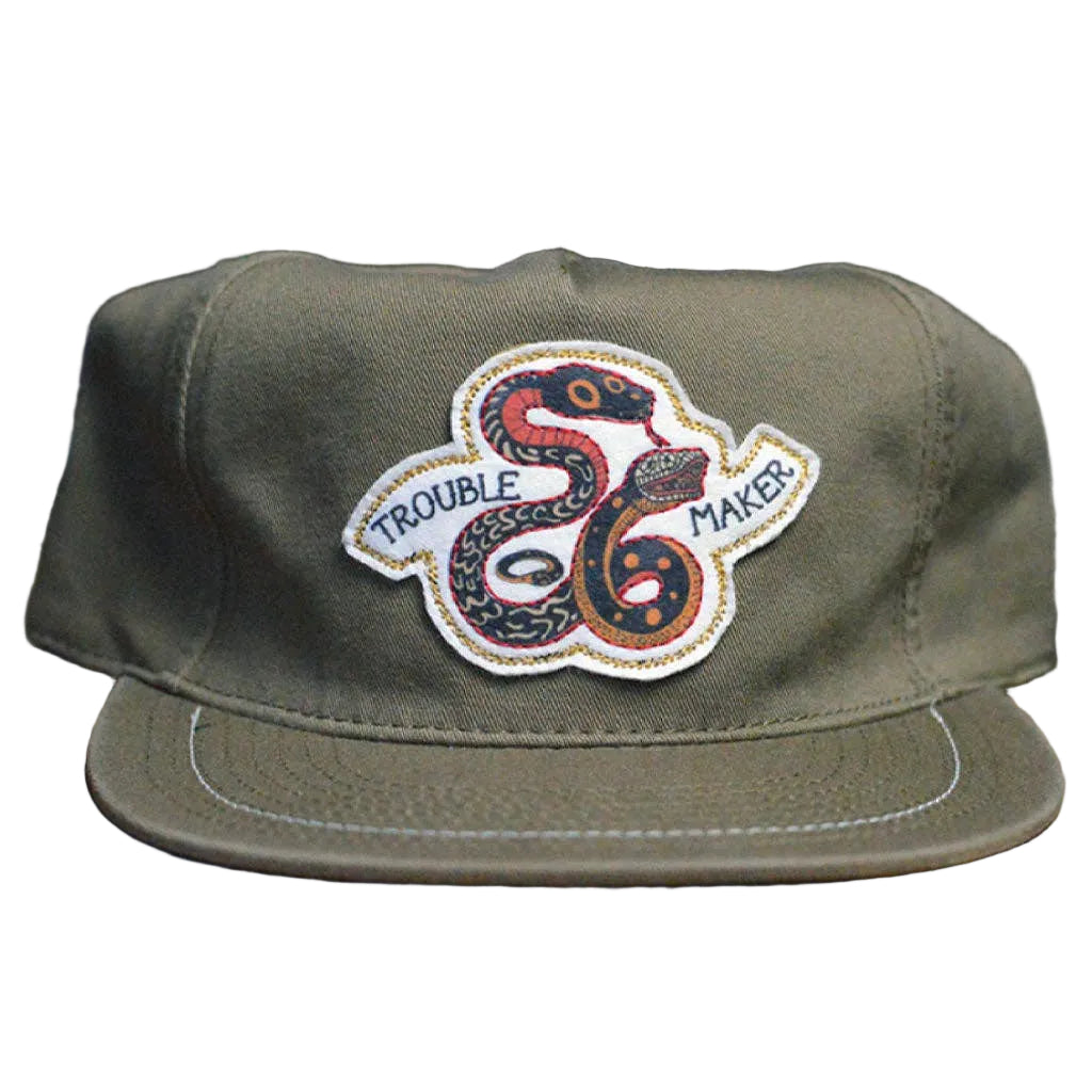Green cap with a snake design and 'Trouble Maker' text on a wooden surface.
