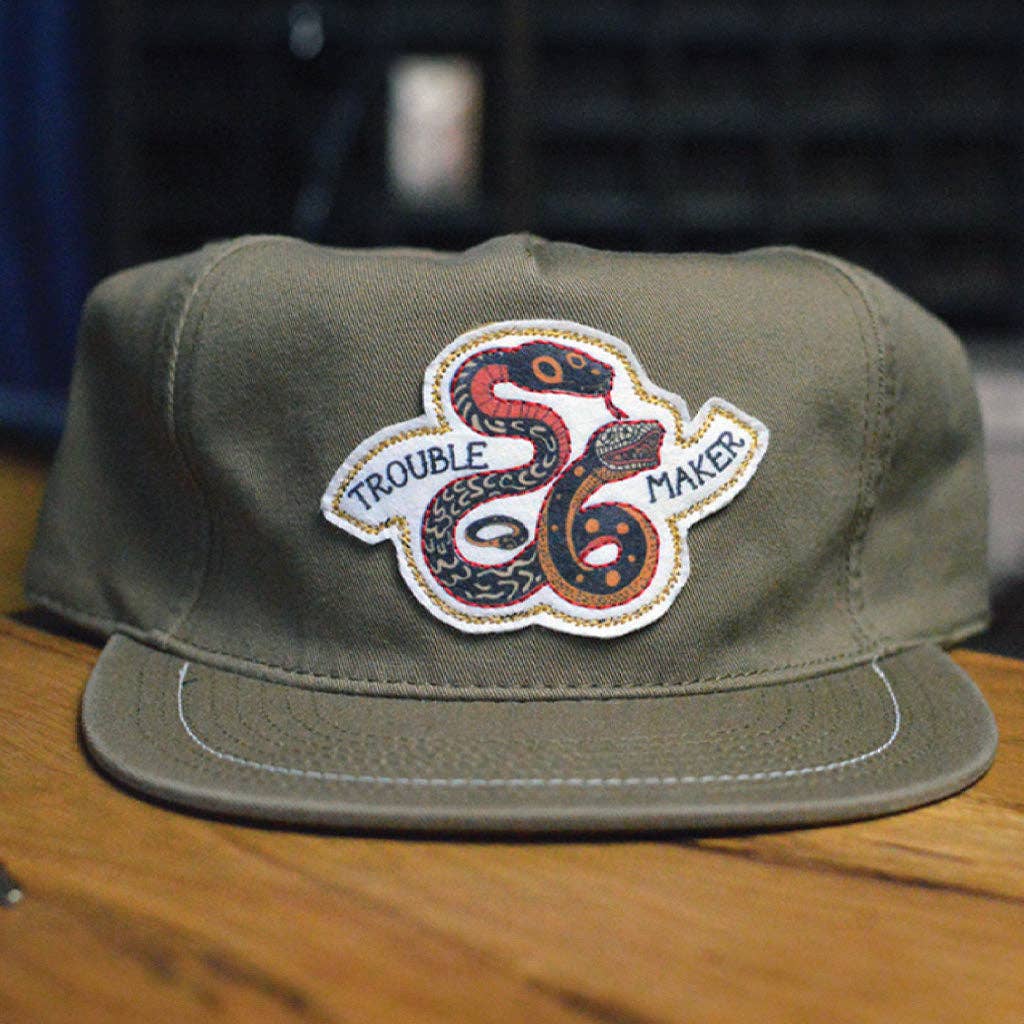 Green cap with a snake design and 'Trouble Maker' text on a wooden surface.