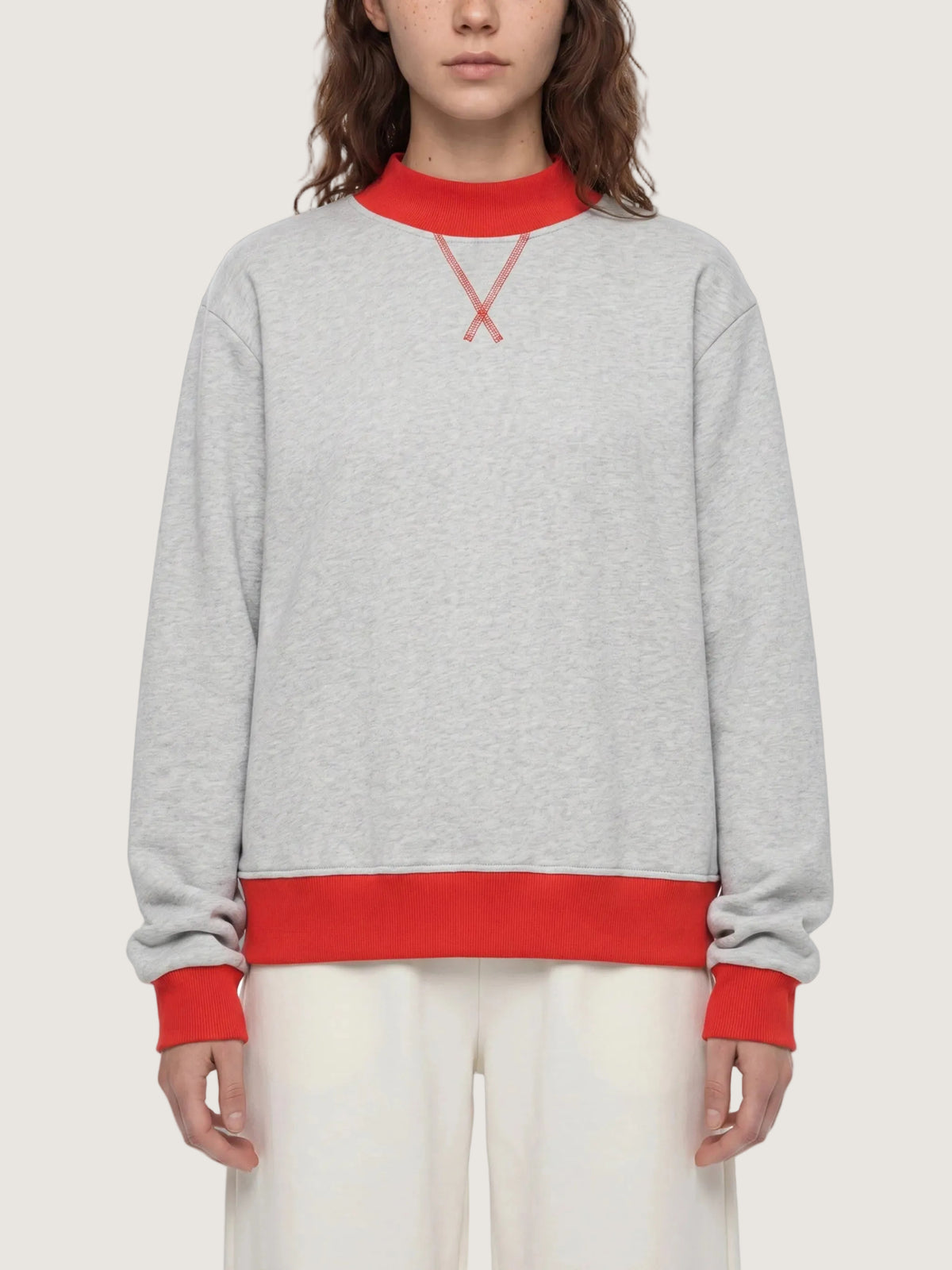 A model wearing a light gray heather crewneck sweatshirt with contrasting red ribbed collar, cuffs, and hem, featuring a red embroidered X logo on the chest.