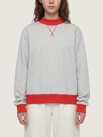 A model wearing a light gray heather crewneck sweatshirt with contrasting red ribbed collar, cuffs, and hem, featuring a red embroidered X logo on the chest.
