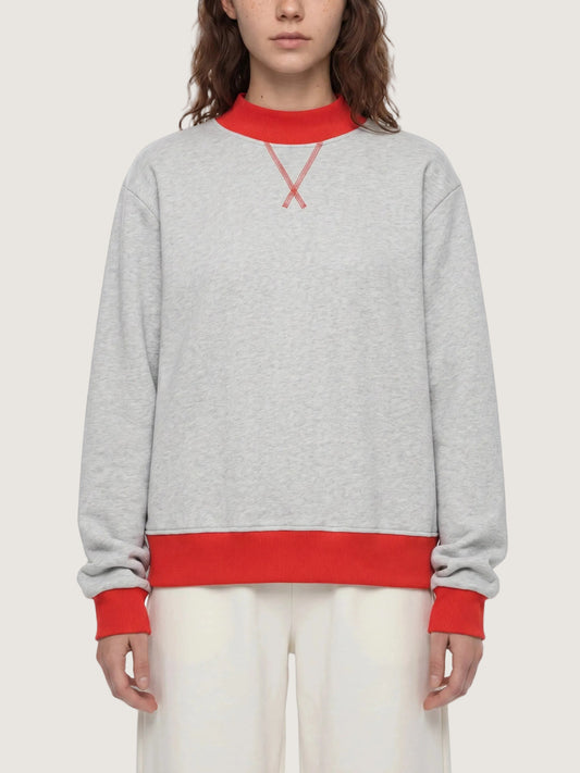 A model wearing a light gray heather crewneck sweatshirt with contrasting red ribbed collar, cuffs, and hem, featuring a red embroidered X logo on the chest.