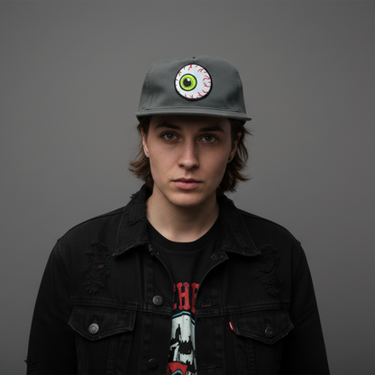 A young man with shoulder-length brown hair stands against a solid dark gray background, wearing a dark gray baseball cap with a circular green and white eye patch graphic, a black denim jacket, and a black graphic t-shirt.